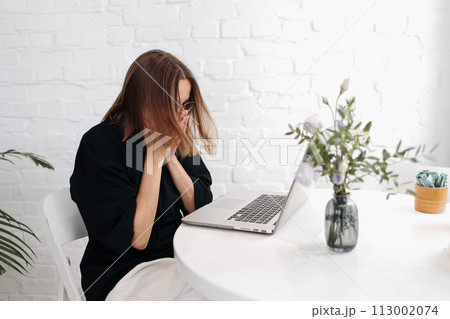 Sick woman sneezes, suffers from seasonal allergy during remote work at laptop by the table at home. Female freelancer or entrepreneur blows nose in office, virus, flu, fever,cold and illness symptom. 113002074