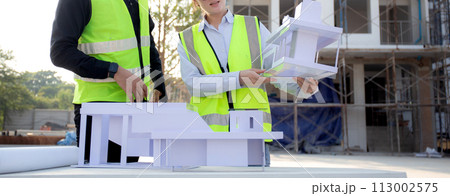 Team architect and builder talking and holding model house at construction site. 113002575