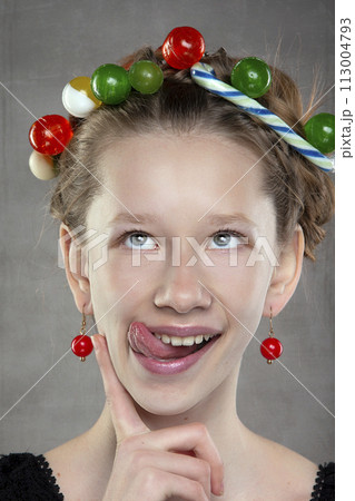 A funny teenager girl with a wreath of sweets on her head shows her tongue. A funny teenager girl with a wreath of sweets on her head shows her tongue. 113004793