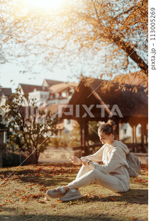 Young Fashionable Teenage Girl With Smartphone In Europian Park In Autumn Sitting At Smiling. Trendy Young Woman In Fall In Park Texting. Retouched, Vibrant Colors. Beautiful Blonde Teenage Girl 113005269