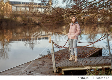 A young woman standing at the shore looking at the river in autumn sunny day. Street view, copy space for text, travel photo. Happy tourist woman on the bank of the river in autumn in warm clothes A young woman standing at the shore looking at the river in autumn sunny day. Street view, copy space for text, travel photo. Happy tourist woman on the bank of the river in autumn in warm clothes 113005352