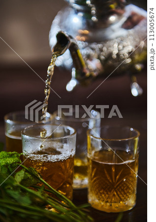 Pouring delicious Moroccan mint tea from a silver teapot into drinking glasses. Bunch of fresh mint in the foreground 113005674