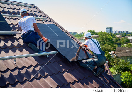 Workers building solar panel system on rooftop of house. Two men installers in helmets installing photovoltaic solar module outdoors. Alternative, green and renewable energy generation concept. 113006532