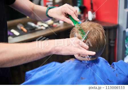 Man barber cutting little boy's hair using comb and scissors. Child getting haircut from adult male, likely barber. Professional hairdresser and cute client at modern barbershop. 113006533
