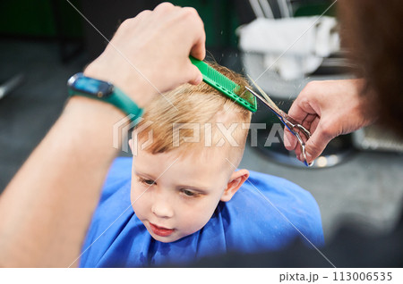 Man barber cutting little boy's hair using comb and scissors. Child getting haircut from adult male, likely barber. Professional hairdresser and cute client at modern barbershop. 113006535