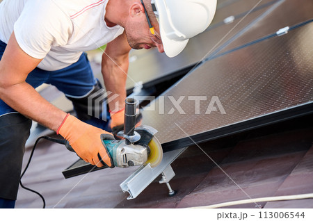 Worker installing or maintaining photovoltaic solar panel system on roof of house. Close up technician using grinder on metal part of the panel's mounting structure. Concept renewable energy. Worker installing or maintaining photovoltaic solar panel system on roof of house. Close up technician using grinder on metal part of the panel's mounting structure. Concept renewable energy. 113006544