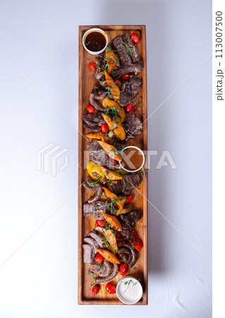 A long wooden platter filled with an assortment of meats and vegetables, including steak, sausage, chicken, potatoes, carrots, and peppers A long wooden platter filled with an assortment of meats and vegetables, including steak, sausage, chicken, potatoes, carrots, and peppers 113007500