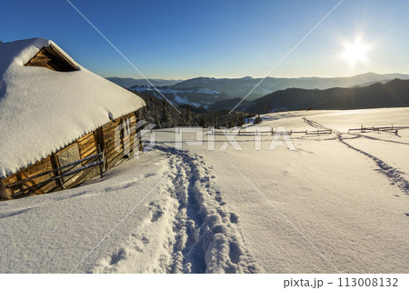 Human footprint path in white deep snow leading to small old wooden forsaken shepherd hut in mountain valley, spruce forest, woody dark hills, bright sun on clear blue sky copy space background. Human footprint path in white deep snow leading to small old wooden forsaken shepherd hut in mountain valley, spruce forest, woody dark hills, bright sun on clear blue sky copy space background. 113008132