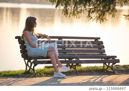 Young woman in summer dress sitting relaxed on lake side bench on warm evening. Wellness resting from everyday rush concept. 113008497