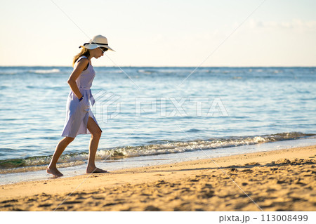 Young woman in straw hat and a dress walking alone on empty sand beach at sea shore. Lonely tourist girl looking at horizon over calm ocean surface on vacation trip. Young woman in straw hat and a dress walking alone on empty sand beach at sea shore. Lonely tourist girl looking at horizon over calm ocean surface on vacation trip. 113008499