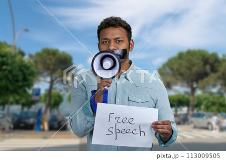 Young man with taped mouth trying to speak into megaphone standing outdoors. Free speech concept. 113009505