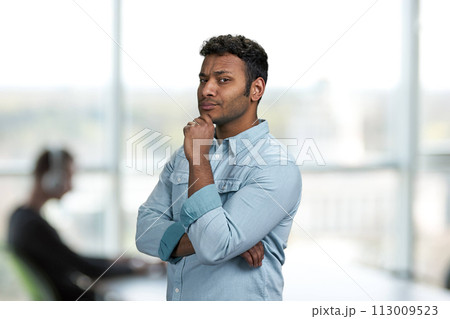 Thoughtful young man holding hand under chin. Office interior blur background. Young man with suspicious expression. Thoughtful young man holding hand under chin. Office interior blur background. Young man with suspicious expression. 113009523