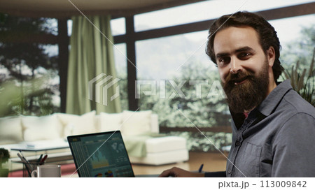 Casual wealthy guy at extravagant holiday residence using laptop to monitor revenue trends for monetary investments and financial growth. Billionaire operating from his lavish forest cabin. 113009842