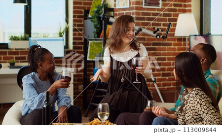 Caucasian woman entertaining friends at house party, sharing with them interesting story. Guest making inclusive group of people laugh in living room while enjoying drinks and charcuterie platter 113009844