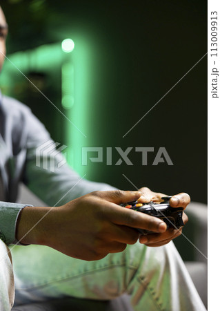 Gamer on couch using controller and headphones to play videogame in neon lit apartment, close up. Player in RGB illuminated living room using high tech gaming console joystick Gamer on couch using controller and headphones to play videogame in neon lit apartment, close up. Player in RGB illuminated living room using high tech gaming console joystick 113009913