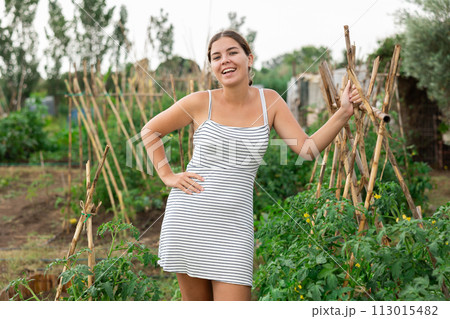 Cheerful organic farmer female holding gloves in her hands stands on her farm. 113015482