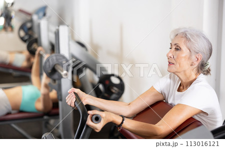 Elderly woman trains her arm muscles on machine Elderly woman trains her arm muscles on machine 113016121