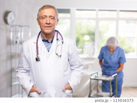 Old man doctor posing in doctor's cabinet with clinical chair Old man doctor posing in doctor's cabinet with clinical chair 113016373