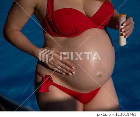 Close-up of the belly of a pregnant woman sunbathing in a red bikini. Expectant mother applies sunscreen.  113019463