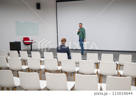 A red-haired Caucasian businesswoman sits in the front row of an empty conference room. Bearded man giving a lecture.  113019844