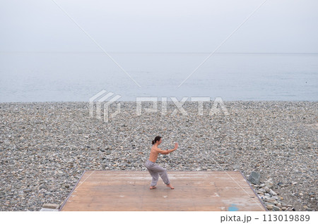 Caucasian man with naked torso practicing wushu on the seashore.  113019889