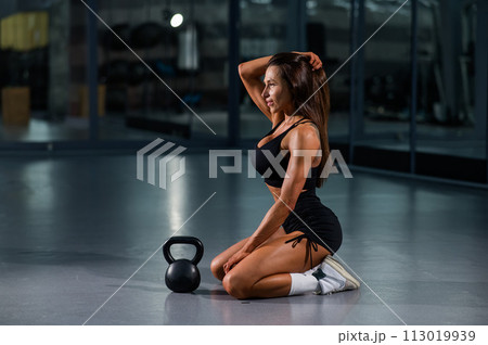 A beautiful brunette with large breasts sits on the floor in the gym. Caucasian woman posing with kettlebell. 113019939