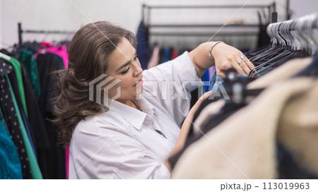 A fat woman in a plus size store chooses clothes while sorting through hangers. A fat woman in a plus size store chooses clothes while sorting through hangers. 113019963