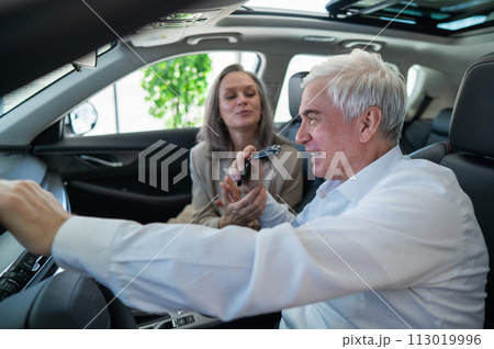 An elderly man holds the keys while sitting in a new car. A gray-haired married couple is happy about buying a car. 113019996