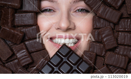 The face of a caucasian woman surrounded by chocolates. The girl eats a bar of chocolate. 113020019