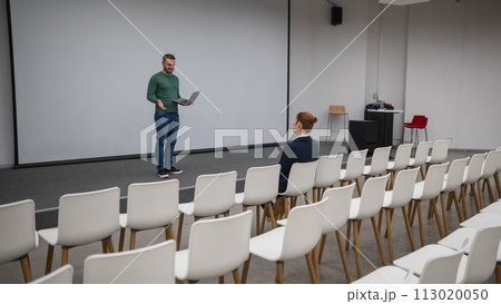 A red-haired Caucasian businesswoman sits in the front row of an empty conference room. Bearded man giving a lecture. A red-haired Caucasian businesswoman sits in the front row of an empty conference room. Bearded man giving a lecture. 113020050