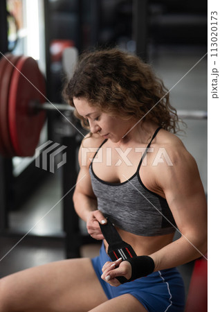 Caucasian forty-year-old woman puts on wrist straps before doing barbell exercises in the gym. Vertical photo.  113020173