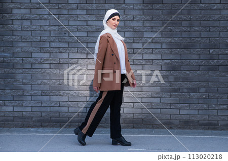 A young woman dressed in a hijab and a business suit walks along a brick wall.  113020218