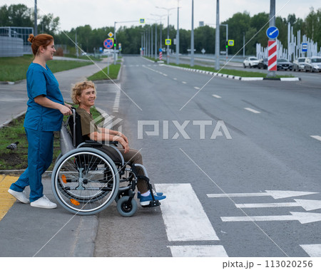 Red-haired nurse pushing an elderly woman in a wheelchair across the road. Red-haired nurse pushing an elderly woman in a wheelchair across the road. 113020256