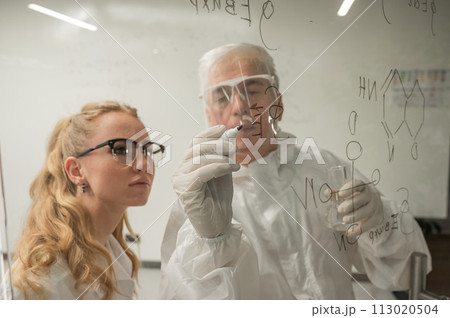 Two chemist colleagues write formulas on glass. Caucasian elderly man and young woman brainstorming. Two chemist colleagues write formulas on glass. Caucasian elderly man and young woman brainstorming. 113020504