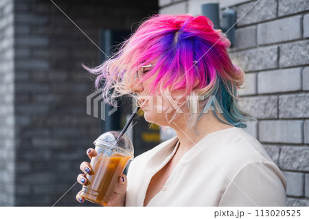 Close-up portrait of curly Caucasian woman with multi-colored hair wearing glasses. The hairstyle model is drinking a cold drink Close-up portrait of curly Caucasian woman with multi-colored hair wearing glasses. The hairstyle model is drinking a cold drink 113020525