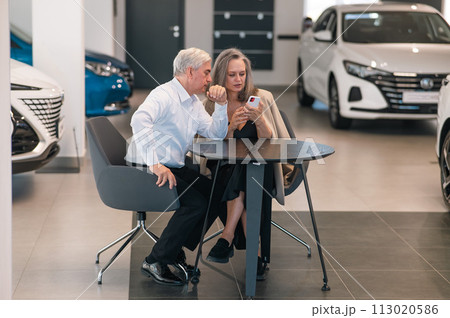 A mature couple is sitting in a car dealership and looking at a smartphone.  113020586