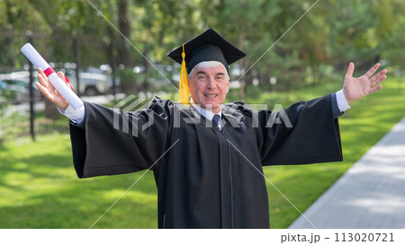 An elderly gray-haired man in a graduate robe spread his arms to the sides and holds a diploma outdoors. An elderly gray-haired man in a graduate robe spread his arms to the sides and holds a diploma outdoors. 113020721