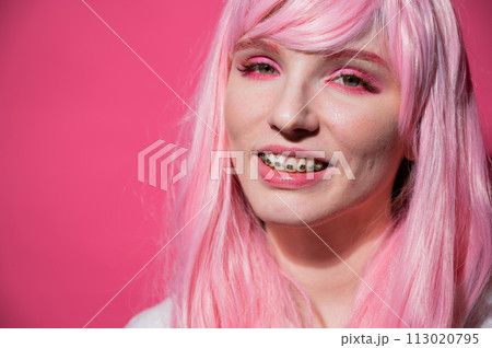 Close-up portrait of a young woman with braces in a pink wig on a pink background. Close-up portrait of a young woman with braces in a pink wig on a pink background. 113020795