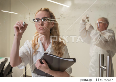 A woman chemist writes a formula on glass. An elderly Caucasian man in a protective suit is doing tests. A woman chemist writes a formula on glass. An elderly Caucasian man in a protective suit is doing tests. 113020987