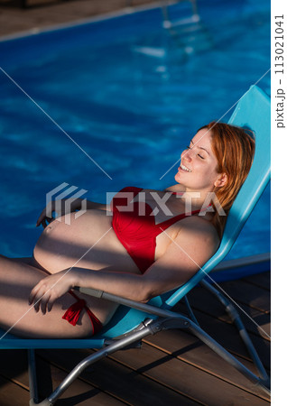 Red-haired pregnant woman in a red bikini sunbathes lying on a sun lounger next to the pool. Red-haired pregnant woman in a red bikini sunbathes lying on a sun lounger next to the pool. 113021041