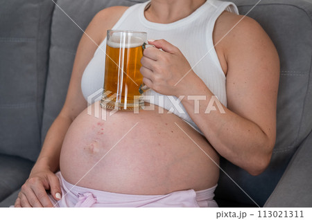 A faceless pregnant woman sits on the sofa and holds a glass of beer on her stomach. Skin rash.  113021311