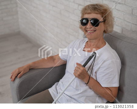 An elderly blind woman wearing sunglasses and with a tactile cane sits on the sofa. An elderly blind woman wearing sunglasses and with a tactile cane sits on the sofa. 113021358