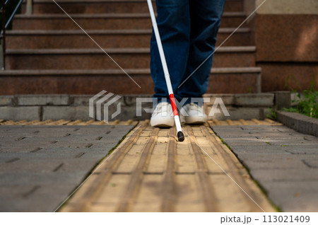 A blind woman walks outdoors using a cane along a tactile yellow tile. 113021409
