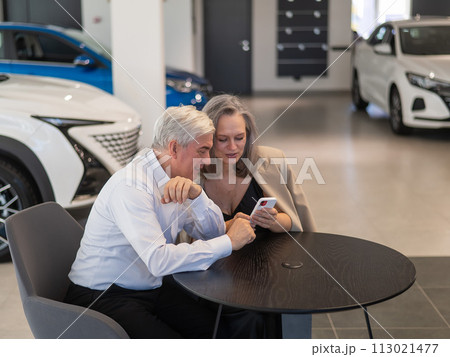 A mature couple is sitting in a car dealership and looking at a smartphone.  113021477