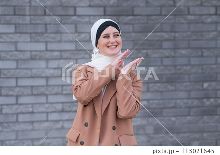 Portrait of a young blue-eyed woman in a hijab against a gray brick wall. Muslim woman happily clapping her hands.  113021645