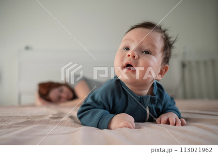 A three-month-old boy lies on his stomach on the bed and his mother sleeps behind him. Postpartum depression. A three-month-old boy lies on his stomach on the bed and his mother sleeps behind him. Postpartum depression. 113021962