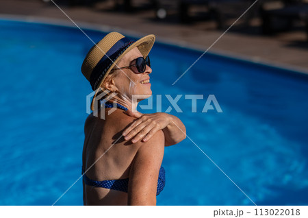 Portrait of an old woman in a straw hat, sunglasses and a swimsuit applying sunscreen to her skin while relaxing by the pool. Portrait of an old woman in a straw hat, sunglasses and a swimsuit applying sunscreen to her skin while relaxing by the pool. 113022018