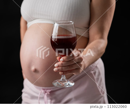 Close-up of the belly of a pregnant woman holding a glass of red wine on a black background.  113022060