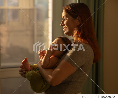 Caucasian woman tenderly holds her newborn son while standing near the window. Caucasian woman tenderly holds her newborn son while standing near the window. 113022072