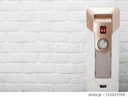 Modern electric oil heating radiator against a white brick wall. The concept of turning on heating in apartments. The cold season is autumn and winter. Copy space for text Modern electric oil heating radiator against a white brick wall. The concept of turning on heating in apartments. The cold season is autumn and winter. Copy space for text 113024769